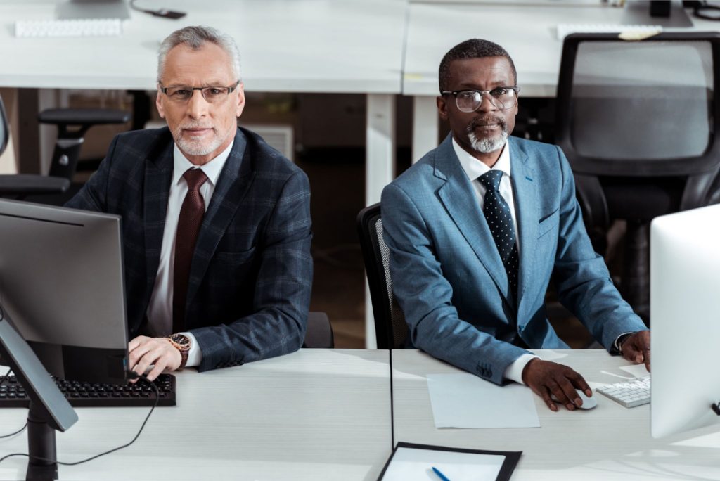 two business partners sitting at desk with computer monitors looking at camera